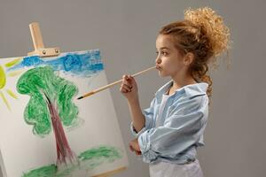 charmant school- meisje is schilderij met een waterverf borstel Aan een ezel, staand Aan een grijs achtergrond. foto