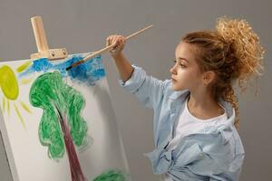 charmant school- meisje is schilderij met een waterverf borstel Aan een ezel, staand Aan een grijs achtergrond. foto