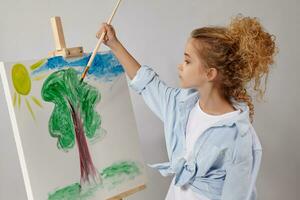 charmant school- meisje is schilderij met een waterverf borstel Aan een ezel, staand Aan een grijs achtergrond. foto