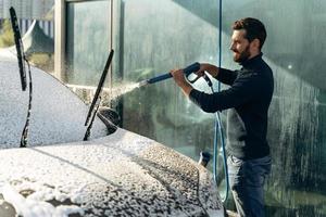 auto wassen. alleen autowassen op de speciale plek. man die lacht naar de camera tijdens het wassen van zwarte auto. auto schoonmaken met behulp van hogedrukwaterconcept foto