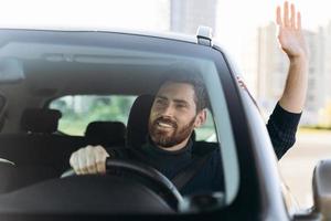 Hallo zeggen. knappe jonge glimlachende zakenman die in een nieuwe auto zit en naar iemand zwaait tijdens het autorijden met plezieremoties foto