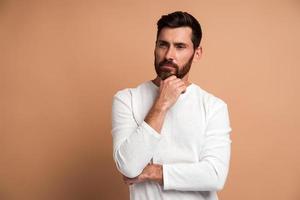 portret van een peinzende knappe bebaarde jonge man die staat, zijn gezicht aanraakt, opzij kijkt en aan iets denkt. indoor studio-opname, geïsoleerd op beige achtergrond foto