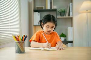 Aziatisch baby meisje kind glimlachen concentreren schrijven tekening gekleurde potlood Aan Notitie boek studie online Aan hout tafel bureau in leven kamer Bij huis. onderwijs aan het leren online van huis concept. foto