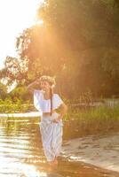 jong mooi Kaukasisch vrouw staand Bij de bank van rivier. traditioneel platteland afbeelding met meisje Bij voorgrond en kopiëren ruimte. zon gloed foto