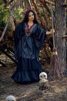 heks in zwart, lang jurk, met rood kroon in haar lang haar. poseren met bezem en schedel in pijnboom Woud. spreuken, magie en hekserij. vol lengte. foto