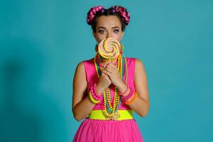 lief meisje met een veelkleurig vlechtjes kapsel en helder verzinnen, poseren in studio tegen een blauw achtergrond, Holding een lolly in haar hand. foto