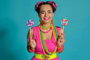 lief meisje met een veelkleurig vlechtjes kapsel en helder verzinnen, poseren in studio tegen een blauw achtergrond, Holding een lolly in haar hand. foto