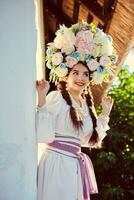 brunette meisje in een wit oekraïens authentiek nationaal kostuum en een krans van bloemen is poseren tegen een wit hut. foto