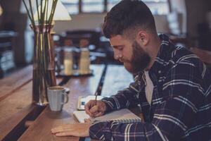 jong gebaard zakenman zit in cafe, huis Bij tafel en schrijft foto