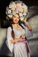 brunette meisje in een wit oekraïens authentiek nationaal kostuum en een krans van bloemen is poseren tegen een wit hut. detailopname. foto