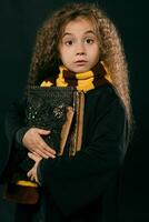 portret van een weinig heks meisje met lang bruin haar- gekleed in donker jas, Holding magie boeken in haar hand, poseren Aan zwart studio achtergrond. foto
