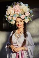 brunette meisje in een wit oekraïens authentiek nationaal kostuum en een krans van bloemen is poseren tegen een wit hut. detailopname. foto