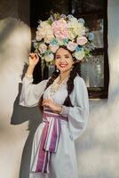 brunette meisje in een wit oekraïens authentiek nationaal kostuum en een krans van bloemen is poseren tegen een wit hut. detailopname. foto