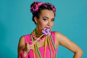 lief meisje met een veelkleurig vlechtjes kapsel en helder verzinnen, poseren in studio tegen een blauw achtergrond, Holding een lolly in haar hand. foto