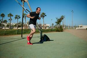 vastbesloten mannetje atleet het uitvoeren van lichaam gewicht opleiding, aan het doen lunges met suspensie riemen Aan buitenshuis sport- grond foto