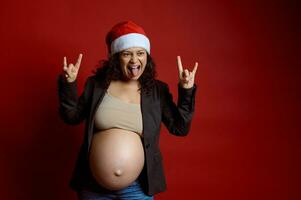 charismatisch emotioneel zwanger vrouw grimassen, staand geïsoleerd over- rood kleur achtergrond, gekleed in de kerstman hoed foto