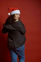 terug visie mooi etnisch jong volwassen zwanger vrouw vervelend de kerstman hoed, glimlacht schattig op zoek Bij camera, rood achtergrond foto