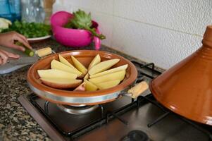 een klei schotel Aan de fornuis, met plakjes aardappel Aan de keuken balie. vrouw Koken Marokkaans tajine foto