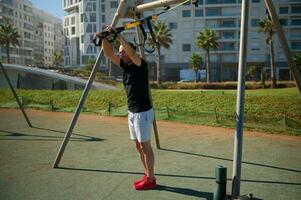 mannetje atleet aan het doen Duwen ups oefening met suspensie riemen, genieten van een ochtend- training in de stedelijk sport- grond foto