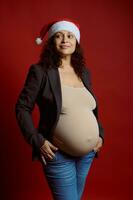 studio portret van emotioneel charismatisch zwanger vrouw in de kerstman hoed, glimlachen op zoek terzijde, geïsoleerd Aan rood achtergrond foto
