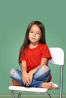 weinig meisje vervelend rood t-kort en poseren Aan stoel foto