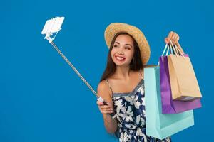 jong elegant vrouw poseren en nemen een selfie Aan de telefoon met boodschappen doen Tassen Aan een blauw achtergrond foto