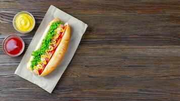 eigengemaakt heet hond met mosterd, ketchup, tomaat en vers salade bladeren Aan bruin houten achtergrond foto