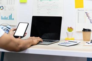 man die een smartphone met een leeg scherm vasthoudt en een laptop, rekenmachine en koffiemok gebruikt met kantoorbenodigdheden op het bureau, gegevensgrafieken op de kantoormuur. foto