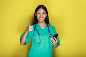 portret van een mooi jong vrouw in een geel achtergrond, Aziatisch vrouw poses met een cel telefoon terwijl vervelend een dokter uniform en een stethoscoop. foto