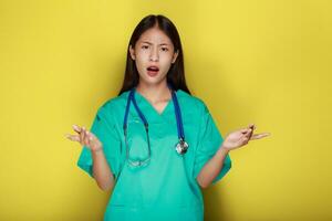 een Aziatisch vrouw vervelend een dokter uniform poses met handen terwijl uitleggen, staand in voorkant van een geel achtergrond. foto