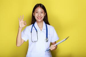 portret van een mooi jong vrouw in een geel achtergrond, mooi Aziatisch vrouw staand gelukkig maken OK gebaar met handen en vervelend dokter uniform. foto