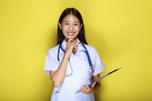 portret van een mooi jong vrouw in een geel achtergrond, Aziatisch vrouw vervelend een dokter uniform maakt een glimlachen uitdrukking, een jong vrouw vervelend een medisch stethoscoop heeft een vriendelijk uitdrukking. foto