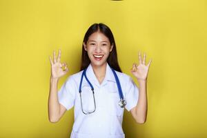 portret van een mooi jong vrouw in een geel achtergrond, mooi Aziatisch vrouw staand gelukkig maken OK gebaar met handen en vervelend dokter uniform. foto