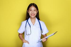 portret van een mooi jong vrouw in een geel achtergrond, Aziatisch vrouw vervelend een dokter uniform maakt een glimlachen uitdrukking, een jong vrouw vervelend een medisch stethoscoop heeft een vriendelijk uitdrukking. foto