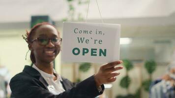 Afrikaanse Amerikaans werknemer opening kleding op te slaan in de ochtend, aan het wachten voor klanten. bipoc kleinhandel arbeider draaien Gesloten bericht teken Aan mode winkel deur in boodschappen doen centrum foto