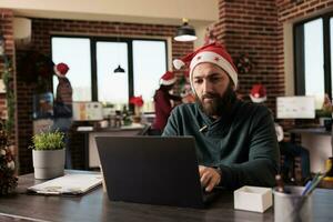 bedrijf werknemer vervelend de kerstman hoed werken Aan laptop in Kerstmis vakantie zakelijke omgeving. Kaukasisch Mens arbeider zittend Bij werkplaats bureau in versierd kantoor Bij Kerstmis vooravond foto