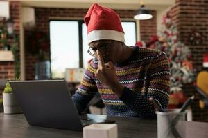 Afrikaanse Amerikaans zakelijke arbeider vaststelling bril terwijl werken Aan laptop in feestelijk versierd kantoor gedurende Kerstmis vakantie. jong Mens vervelend de kerstman hoed beheren taak deadline foto