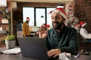 glimlachen werknemer in de kerstman hoed golvend Hoi terwijl sprekend Bij online zakelijke vergadering gebruik makend van laptop. Kaukasisch Mens groet collega's in video-oproep en werken in kantoor Bij Kerstmis vooravond foto