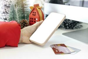 teder handen wieg een smartphone, leeg scherm door feestelijk tafereel een mooi Kerstmis boom versierd met credit kaarten, langs met Kerstmis ballen, pijnboom kegels, en rood huis geschenk doos gebonden gouden lintje. foto