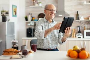 gelukkig senior Mens zoeken Aan tablet in keuken gedurende ontbijt genieten van Loisire tijd. ouderen gepensioneerd persoon werken van huis, telewerken gebruik makend van afgelegen internet baan online communicatie. foto