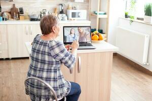 gelukkig senior vrouw gedurende een video conferentie met familie gebruik makend van laptop in keuken. online telefoontje met dochter en nicht. oud ouderen persoon gebruik makend van modern communicatie online internet web technologie. foto