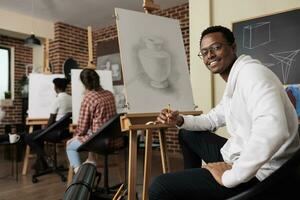 gelukkig Afrikaanse Amerikaans vent zittend Bij ezel in klas genieten van maken kunst gedurende groep tekening klas, creatief ruimte voor sociaal verbinding. glimlachen Mens aan het leren hoe naar trek Bij kunst werkplaats foto