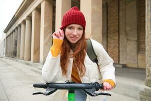 glimlachen roodharige Europese meisje schijven openbaar escooter, toerist onderzoekt stad, ritten in stad centrum foto