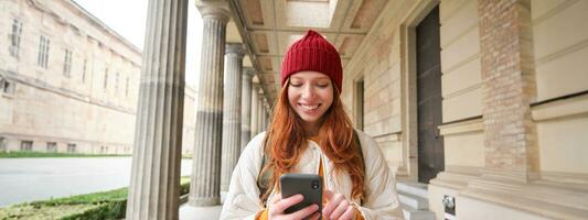 glimlachen vrouw met rood haar, toerist gebruik makend van mobiel telefoon toepassing terwijl wandelen in de omgeving van dorp, zoeken voor attractie naar op bezoek komen, verkennen bezienswaardigheden bekijken met smartphone app foto