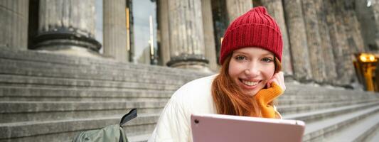 dichtbij omhoog portret van schattig roodharige Europese meisje, zittend in rood hoed in de buurt gemeentelijk gebouw Aan trap, houdt digitaal tablet, leest ebook of surft internet foto