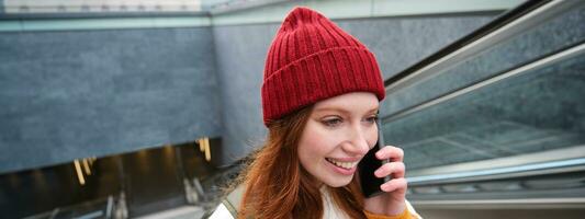 portret van gelukkig roodharige vrouw wandelen in de omgeving van stad- met smartphone, roeping iemand, pratend Aan mobiel telefoon buitenshuis foto