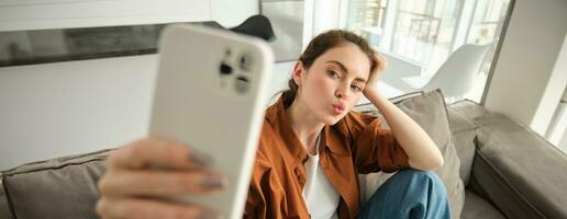 portret van schattig jong vrouw, modern meisje nemen selfies Aan smartphone app, poseren Aan bank, strekt zich uit haar wit mobiel telefoon naar nemen afbeelding, plooi lippen voor foto