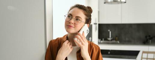 portret van gelukkig vrouw Bij huis, antwoorden telefoon telefoongesprek, pratend Aan mobiel, Holding smartphone en glimlachen foto