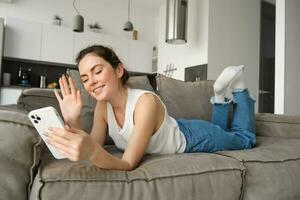 gelukkig schattig vrouw model, aan het liegen Aan bank, golvend hand- Bij smartphone, gezegde hoi, verbindt naar video telefoongesprek, resting Bij huis en pratend naar vriend foto