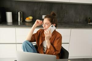 beeld van mooi modern vrouw werken van huis, aan het studeren in keuken met laptop, pratend Aan smartphone, roeping iemand Aan telefoon foto